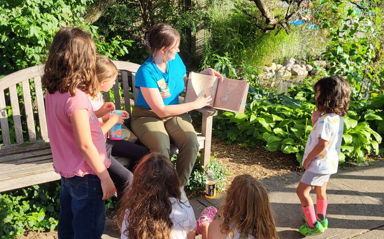 A woman sitting on a bench in a garden reading a book to children.